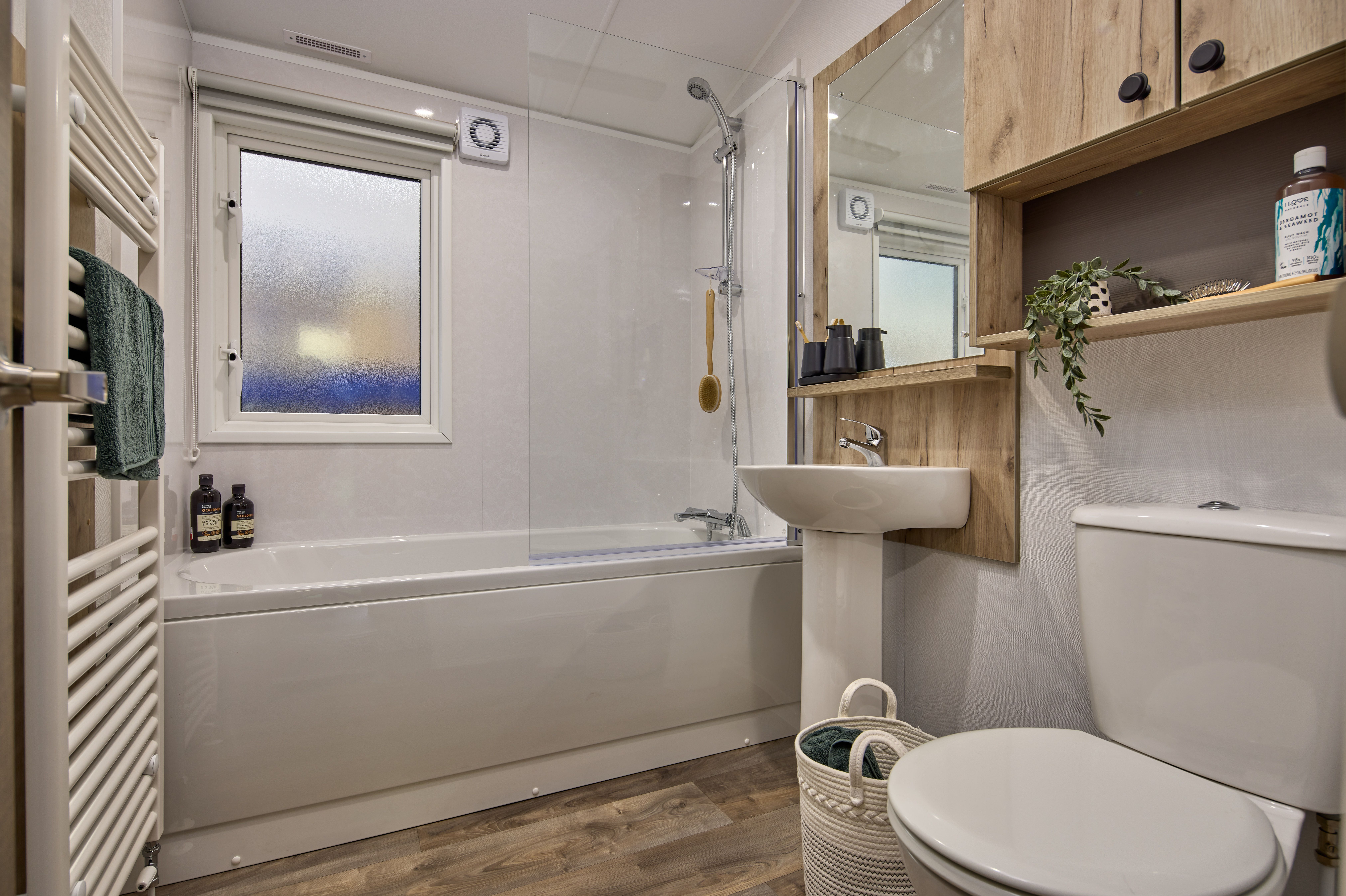 The bathroom in the Willerby Buxton holiday home. There is a large towel rail, shower over bath and big mirror. The white and wood colour scheme makes the most of the light from the large window. 