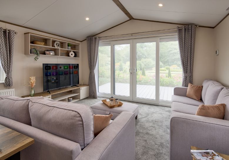 The Willerby Malton Elite spacious lounge with two grey free-standing sofas and a matching footstool with a storage media unit in cream and a dark oak effect.