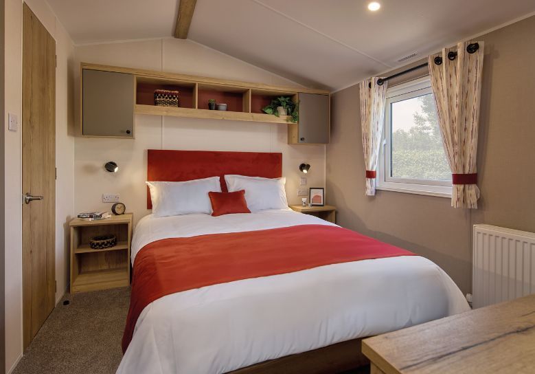 The Willerby Rio master bedroom has a cream accent wall and orange soft furnishings matching the colour scheme, with oak effect storage units around the bed with light grey cupboard doors.