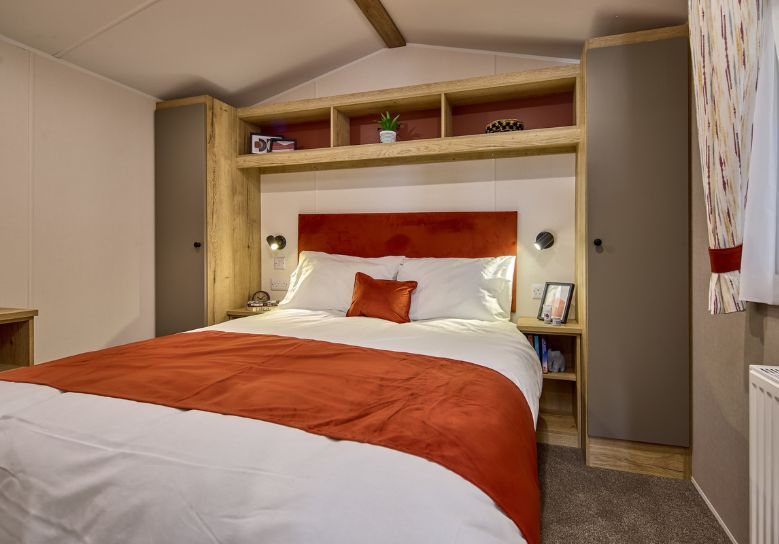 The Willerby Rio Compact master bedroom has a cream accent wall and orange soft furnishings matching the colour scheme, with oak effect storage units around the bed with light grey cupboard doors.