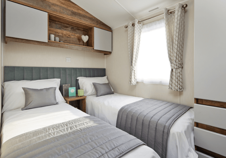 The Willerby Brookwood spacious twin bedroom with overhead storage with white cupboard doors and an upholstered sage green headboard.