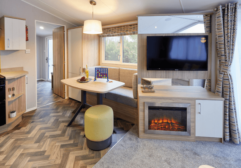 The Willerby Castleton dining area with grey fixed window seat and a yellow stool, with the view of the media unit in the lounge with an optional electric fireplace.