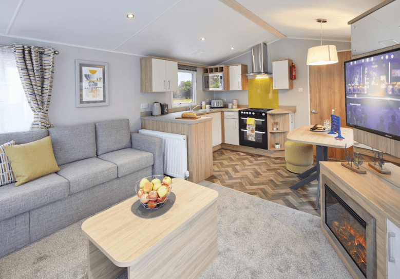 The Willerby Castleton lounge with a grey fixed sofa and a view of the kitchen which has oak effect storage units. 