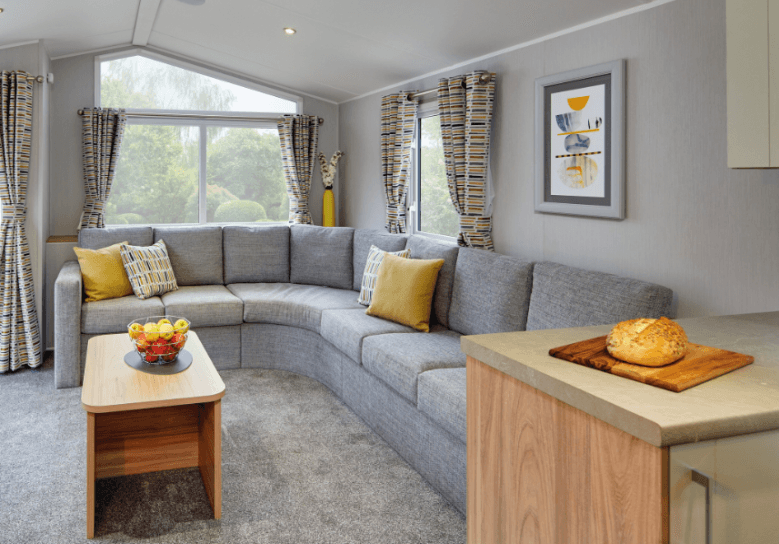 The Willerby Castleton lounge with a grey L-shaped sofa and yellow patterned pillows matching the colour scheme of the home, with a small oak effect coffee table. in the middle of the lounge.