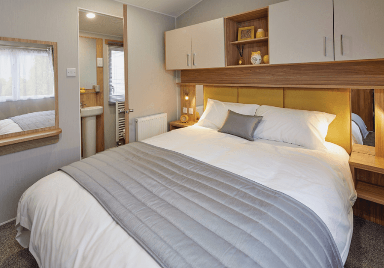 The Willerby Castleton peaceful master bedroom with overhead storage units adorned with white cupboard doors. The bed has a yellow upholstered headboard matching the rest of the home.