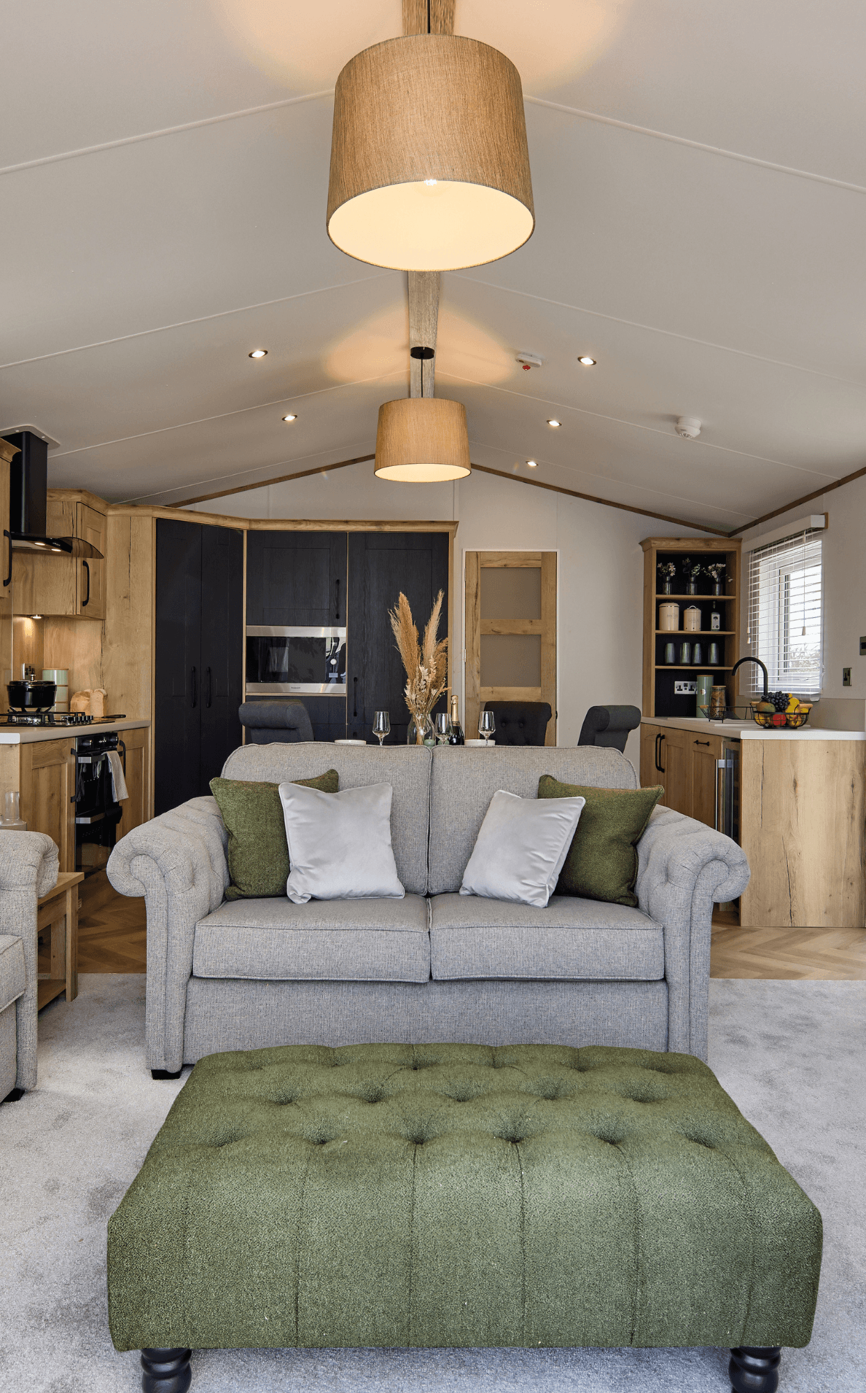 The Willerby Dorchester lounge with grey sofas and a green stylish footstool, with a view of the media unit and kitchen.