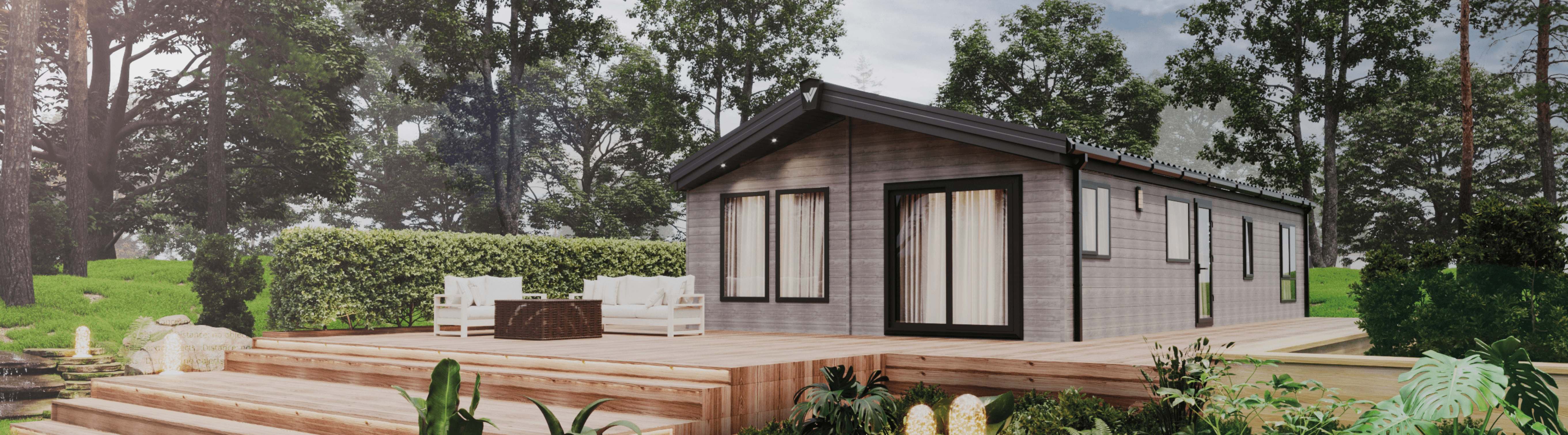 The Willerby New Holland holiday lodge surrounded by trees and plants. There is multi-level decking in front with water features. 