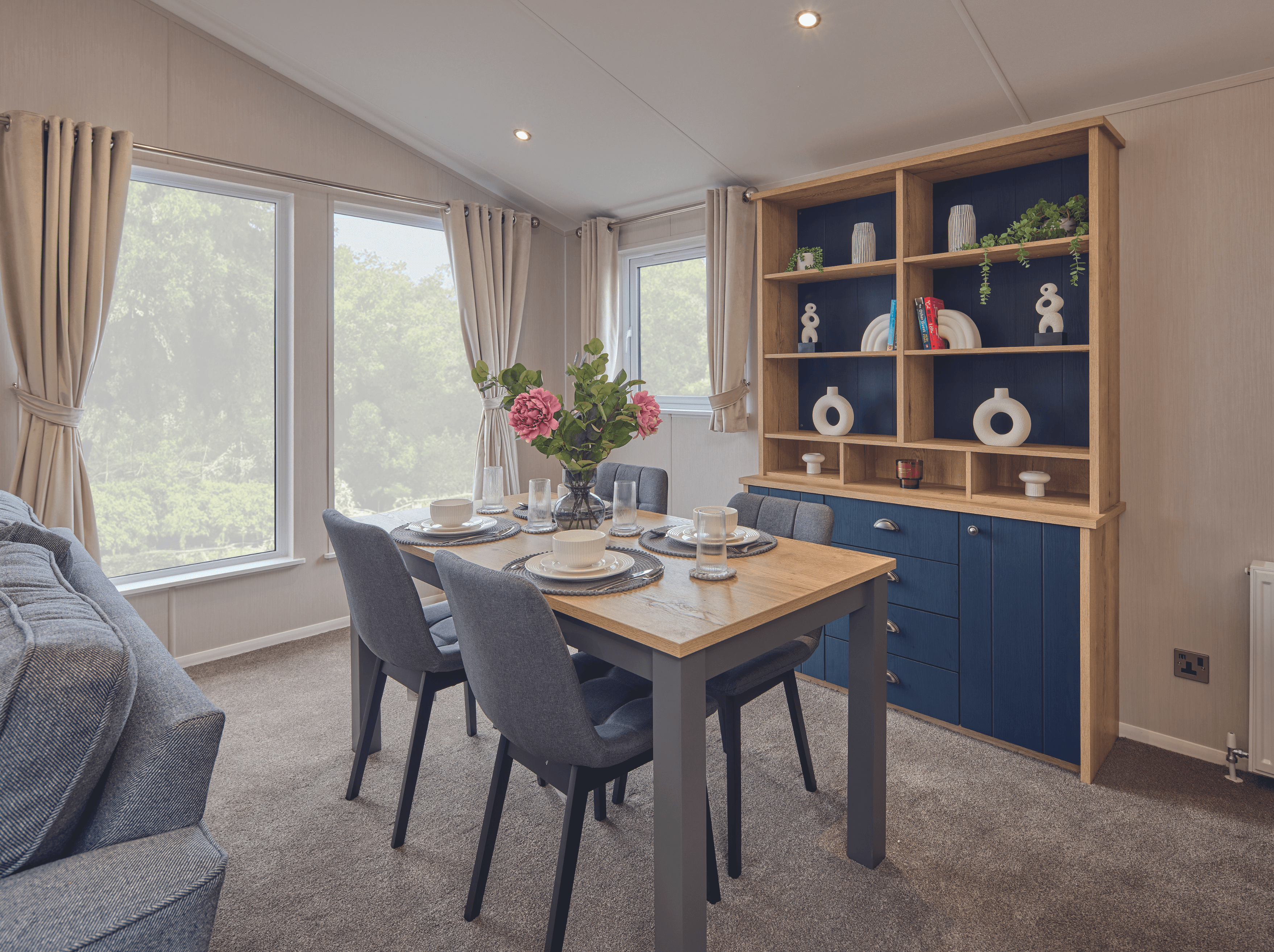 The dining area of the Willerby New Holland holiday lodge. There is a table with seating for four and a fitted dresser against the wall. Full-height windows let in lots of light. 