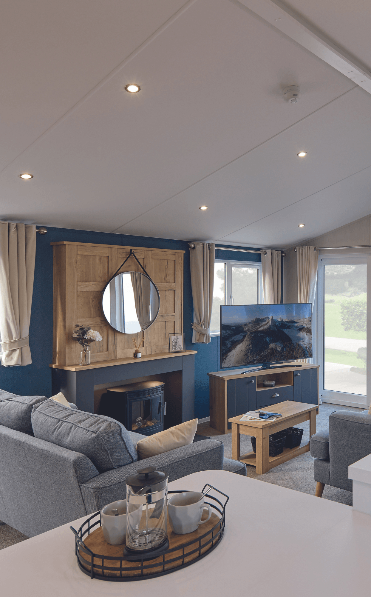 The living area of the Willerby New Holland holiday lodge. There is a large TV on a cabinet, comfortable free-standing sofas, a dining table and breakfast bar. Large full height windows flood the space with light. 