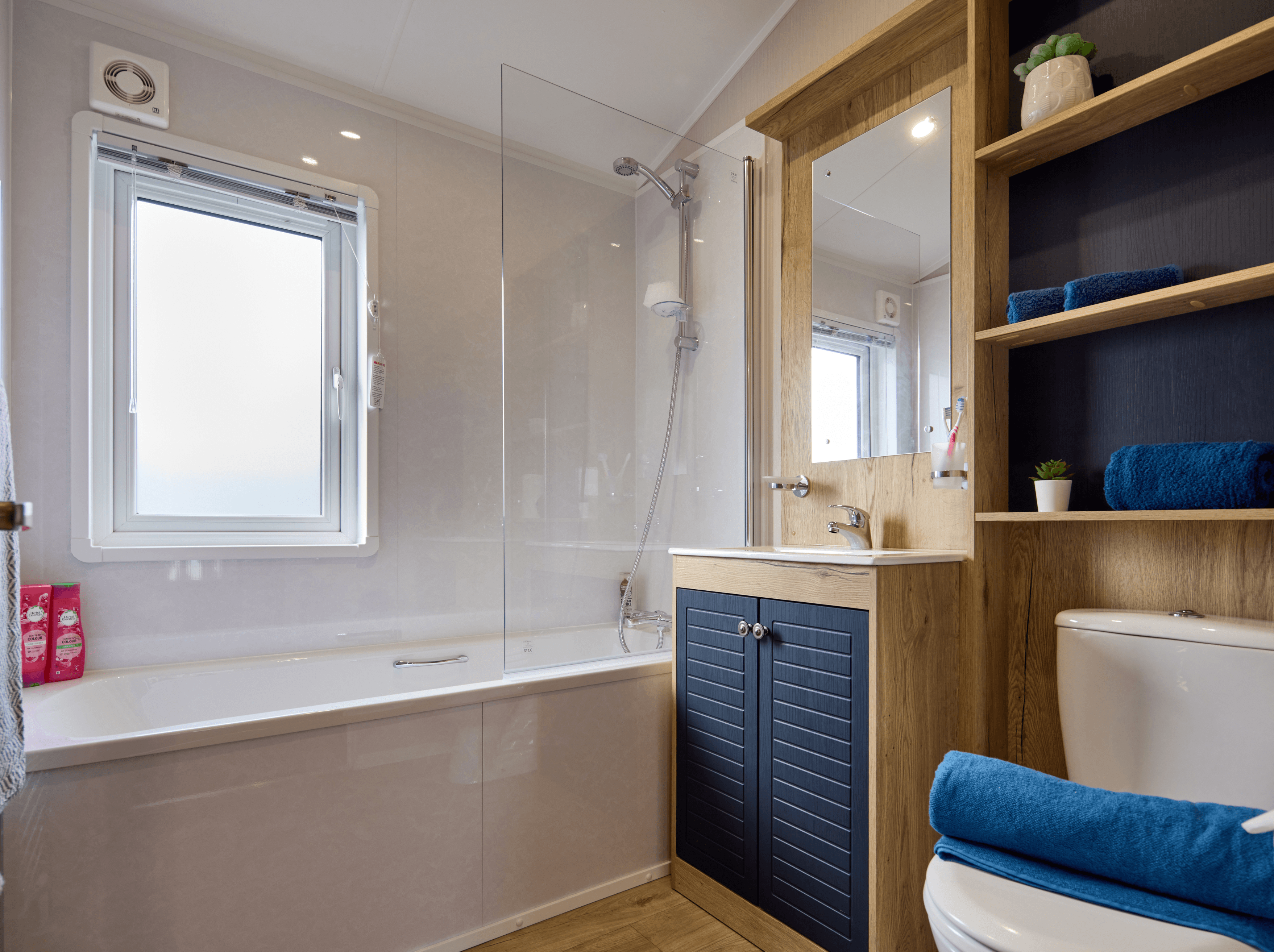 The bathroom in the Willerby New Holland holiday lodge. There is a large bath with shower over and a sink with a large mirror above it. The cupboards and shelves are wood with white walls and dark blue highlights. 