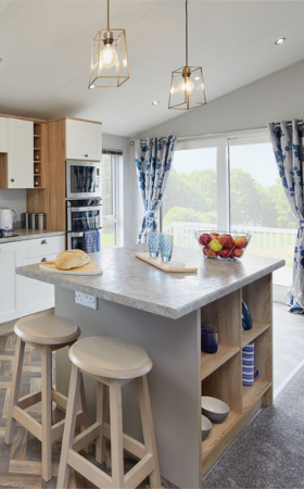 The Willerby Pinehurst kitchen has white cupboard doors with grey worktops and a matching colour scheme island with two oak effect stools.