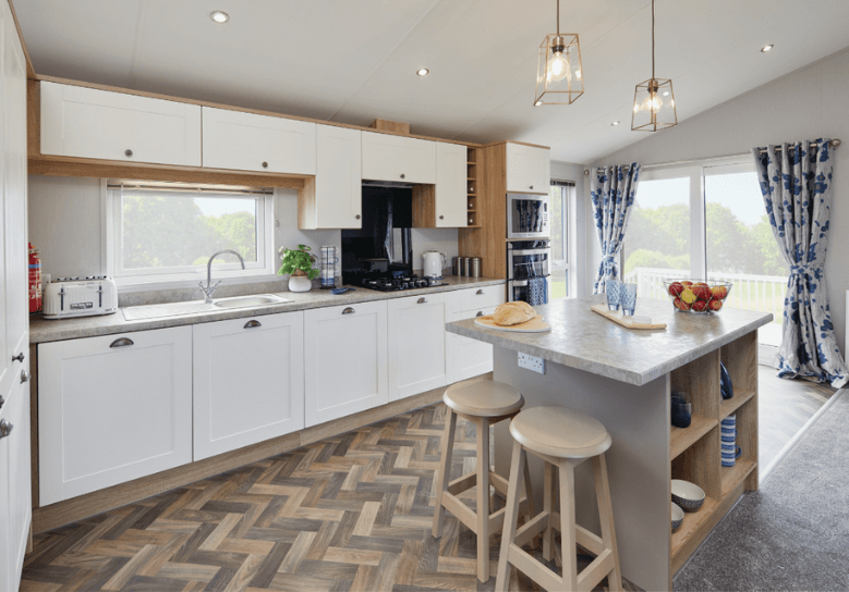 The Willerby Pinehurst kitchen has white cupboard doors with grey worktops and a matching colour scheme island with two oak effect stools.