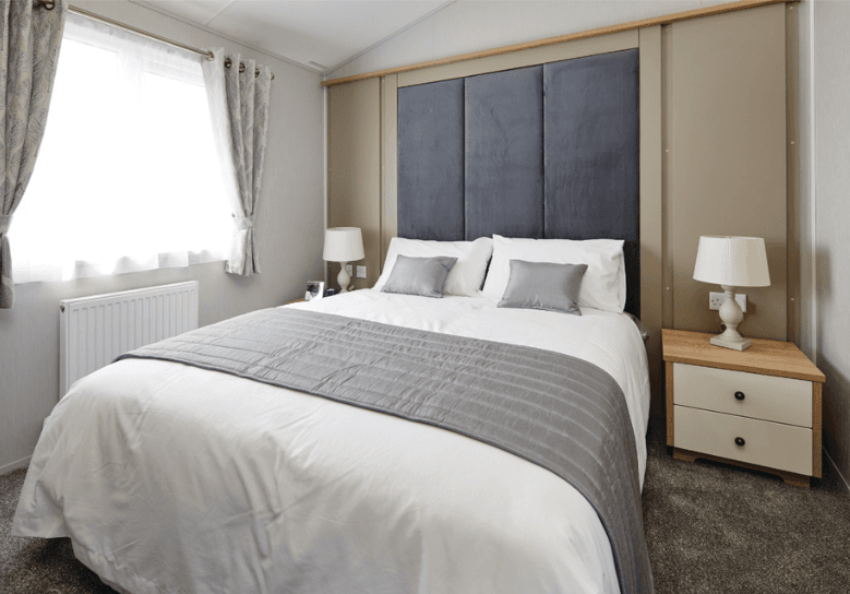 The Willerby Pinehurst master bedroom has a tall grey upholstered headboard on the feature wall, and two oak effect bedside tables with white cupboard doors.