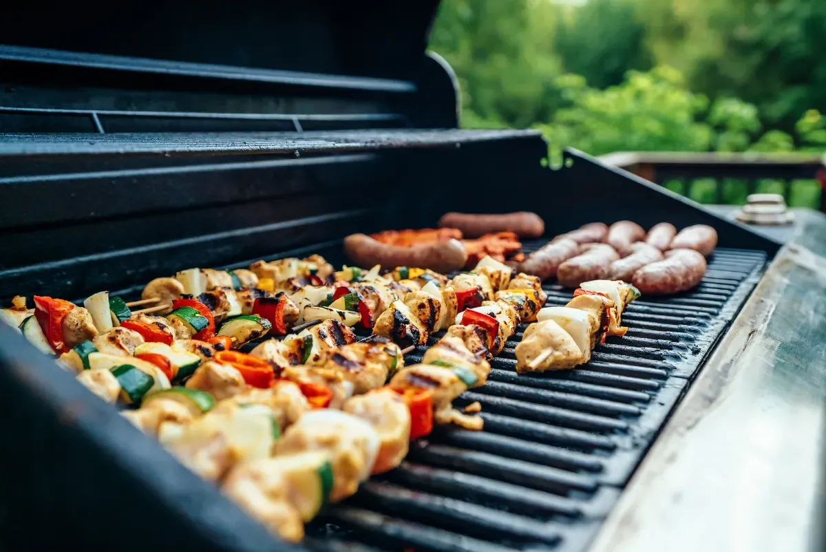 Close-up of a large gas barbecue with blurry green trees in the background. Lots of kebabs, sausages and other food are cooking. 
