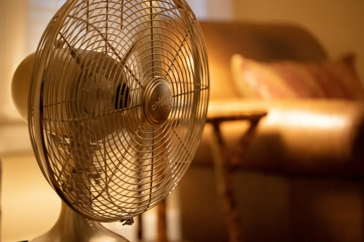 Wire mesh fan blowing cool air through a static caravan. There is a blurry chair and table in the background. 