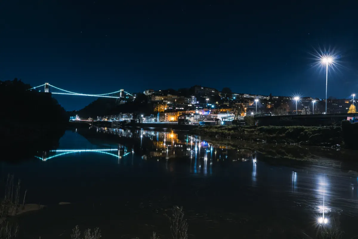 A view of Bristol at night, showing the Clifton Suspension Bridge and Observatory Hill reflected in the River Avon.