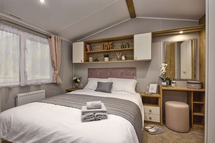 The Willerby Sierra master bedroom has oak effect storage units with white cupboard doors, a matching dressing area and an upholstered pink headboard matching the colour scheme of the home.