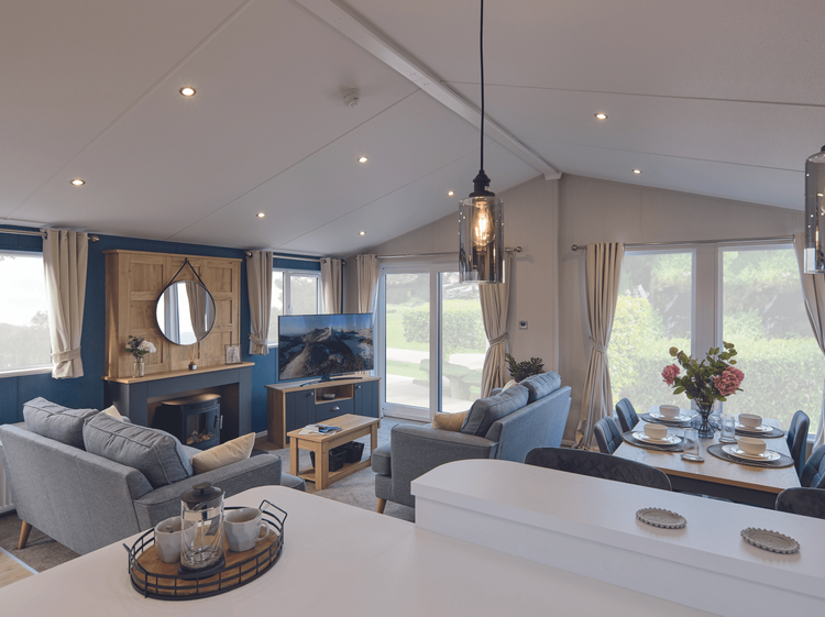 The living area of the Willerby New Holland holiday lodge. There is a large TV on a cabinet, comfortable free-standing sofas, a dining table and breakfast bar. Large full height windows flood the space with light. 