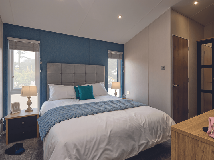 The main bedroom in the Willerby New Holland holiday lodge. The large bed is flanked by two windows and storage units. There is a door leading to the en-suite bathroom. 