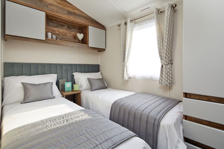 The Willerby Brookwood spacious twin bedroom with overhead storage with white cupboard doors and an upholstered sage green headboard.