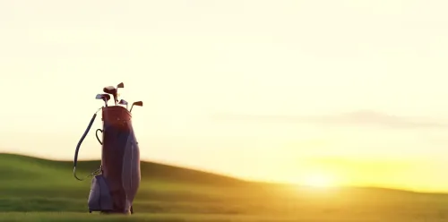 A golf bag full of clubs sits on the fairway bathed in golden light while the sun rises in the background.