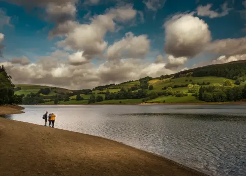 <alt> Three people on staycation on the bank of a reservoir in the Peak District in the UK, looking across to green hills. 