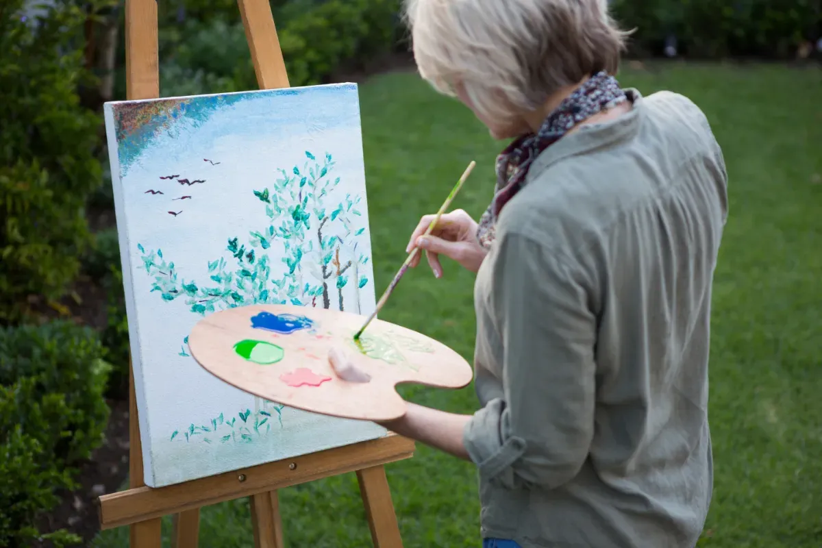 Lady with grey hair painting a landscape on an easel outdoors