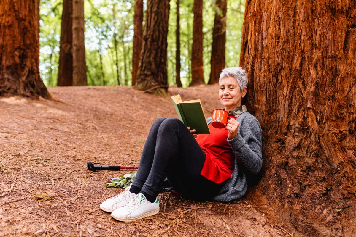 A lady with short, grey hair sitting and reading, leaning against a tree